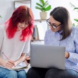 Female teacher teaches teenage girl, sitting together on couch in office, using laptop